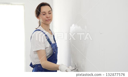 Professional female construction worker wearing blue coveralls is using a finishing trowel to carefully apply plaster on a wall, demonstrating expertise in home renovation 132877576