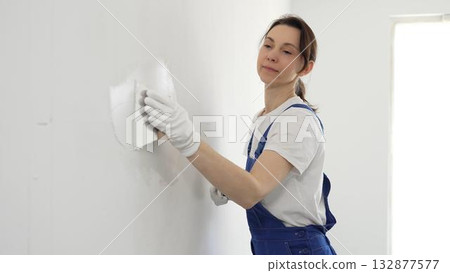 Construction worker woman spreading finishing putty on white wall, wearing protective gloves while smoothing surface with putty knife for precise home repair Construction worker woman spreading finishing putty on white wall, wearing protective gloves while smoothing surface with putty knife for precise home repair 132877577
