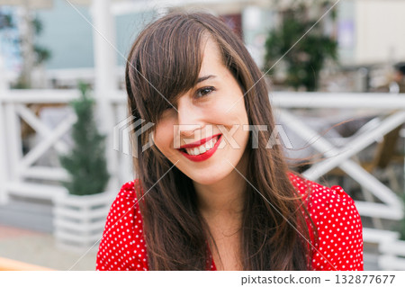 Smiling woman in red polka dot dress outdoors at cafe terrace. Positivity, beauty, and joyful modern lifestyle. 132877677