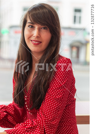 Portrait of a woman in red polka dot dress sitting in cafe and smiling. Confidence, femininity, and natural beauty in modern lifestyle. 132877716