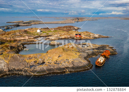 Rusty shipwreck partially submerged in calm coastal waters near a red boathouse on a rocky islet. The Atlantic Ocean Road stretches across Norways wild and rugged seascape Rusty shipwreck partially submerged in calm coastal waters near a red boathouse on a rocky islet. The Atlantic Ocean Road stretches across Norways wild and rugged seascape 132878884