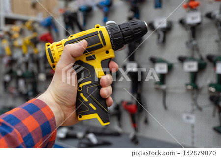 A man in a hardware store chooses a new drill or screwdriver. A hand holds a yellow power drill in front of a hardware store display with various tools. Handyman holding electric screwdriver. A man in a hardware store chooses a new drill or screwdriver. A hand holds a yellow power drill in front of a hardware store display with various tools. Handyman holding electric screwdriver. 132879070