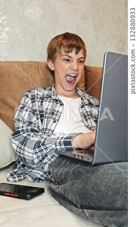 Young man at home smiling with satisfaction, is emotionally working on a laptop. Caucasian guy sitting on sofa and enthusiastically taps on the keyboard. crazy 132880933