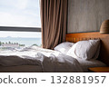 Cozy bed with white sheets and pillow in sunlit room by large window overlooking the sea 132881957