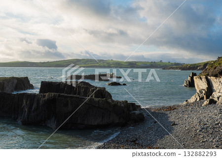 Gentle waves lap against dark rocks and a pebble beach at Sheep Cove. Green hills and a cloudy sky fill the background on a bright day in Ireland. 132882293