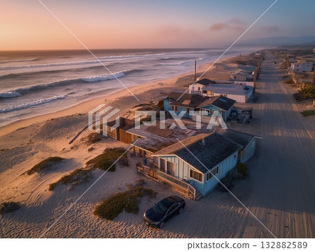 Coastal beach houses at sunset with waves, sandy road and warm pastel sky creating serene seaside atmosphere Coastal beach houses at sunset with waves, sandy road and warm pastel sky creating serene seaside atmosphere 132882589
