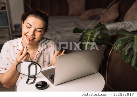 Woman applying lip gloss beside laptop at home 132882917