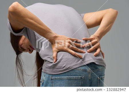 Woman bending forward with hands on lower back, grimacing in pain, studio image Woman bending forward with hands on lower back, grimacing in pain, studio image 132882947