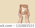 Hands Presenting Christmas Ornament Gift Box With Angel Figurine On Beige Background. Concept Of Holiday Decorations, Festive Packaging Ideas, Christmas Gifting Trends, And Seasonal Home Styling 132883523