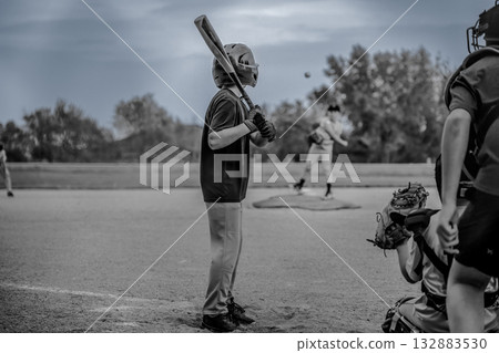 Young player waits at bat as pitcher throws baseball during a game in a local park on a cloudy afternoon Young player waits at bat as pitcher throws baseball during a game in a local park on a cloudy afternoon 132883530