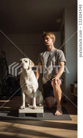 A young man is engaging in squats on a yoga mat, with his Labrador dog standing on a small platform next to him, promoting fitness and companionship in a home setting 132884348
