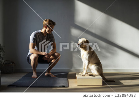 A young man is engaging in squats on a yoga mat, with his Labrador dog standing on a small platform next to him, promoting fitness and companionship in a home setting 132884388