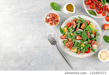 Green salad with prosciutto, arugula, strawberries, melon, tomatoes, parmesan on gray background. Top view. Copy space. Green salad with prosciutto, arugula, strawberries, melon, tomatoes, parmesan on gray background. Top view. Copy space. 132885371
