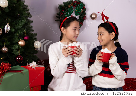girls in Christmas reindeer headbands are eating marshmallows from red mugs. 132885752