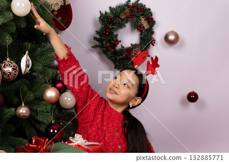 Little girl in red dress decorates Christmas tree with ornaments and lights, Little girl in red dress decorates Christmas tree with ornaments and lights, 132885771