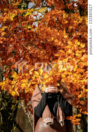 A woman stands near an autumn tree, covering her face with a colorful fall leaf, mid shot, outdoors, seasonal mood 132885827