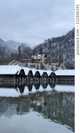 Serene winter landscape with snow-covered boathouses reflecting in calm water surrounded by mountains Serene winter landscape with snow-covered boathouses reflecting in calm water surrounded by mountains 132886343