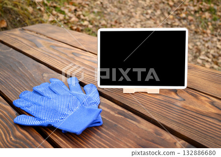 a blue gardening glove and a white tablet on a stand are on a brown wooden table 132886680