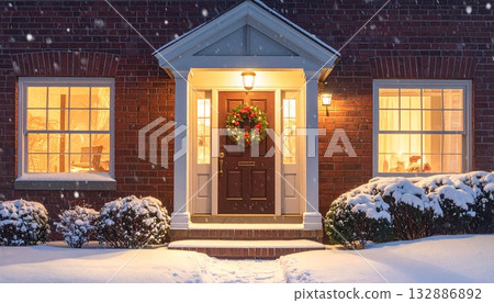 On a snowy night, the brick house and the wreath entrance are lit up with warm lights. 132886892