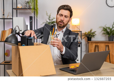 Middle-aged businessman packs personal items in box looks around room with gentle nostalgia softly Middle-aged businessman packs personal items in box looks around room with gentle nostalgia softly 132887203
