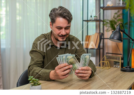 Middle-aged man counts cash smiling showing big financial success dollar bills wealth at table desk 132887219