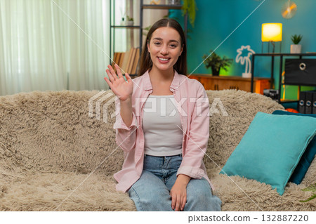 Young woman smiling waving cheerful hello greeting camera with friendly energy on living room sofa 132887220