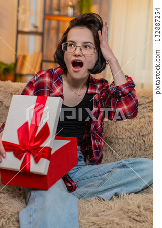 Young happy woman opening gift at home with surprise and joy, reacting with happiness, wide smile Young happy woman opening gift at home with surprise and joy, reacting with happiness, wide smile 132887254