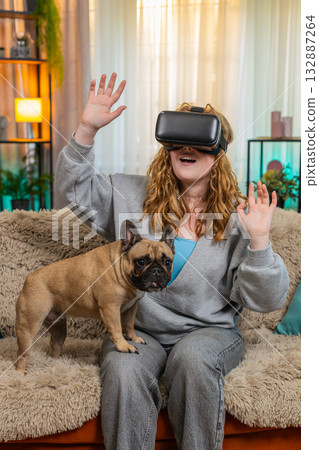 Young woman using VR headset on home sofa with excited movement as pug dog watches with funny face Young woman using VR headset on home sofa with excited movement as pug dog watches with funny face 132887264