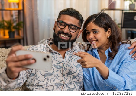Indian family couple man woman laughing together at funny videos on smartphone sitting on home sofa Indian family couple man woman laughing together at funny videos on smartphone sitting on home sofa 132887285