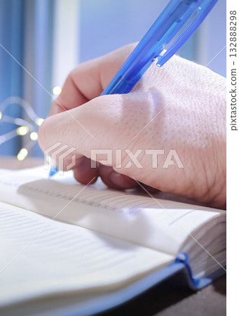 A man writes down his plans and goals for the New Year in a notebook, while small garlands sparkle nearby A man writes down his plans and goals for the New Year in a notebook, while small garlands sparkle nearby 132888298