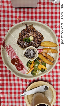 A juicy grilled steak topped with butter and parsley is served with brown sauce, colorful vegetables, and ketchup on a rustic plate, creating a warm and appetizing dining experience full of flavor. 132888902