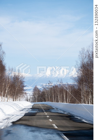 Snow starting to melt on the road and snow-capped mountains of the Tokachi mountain range 132890034