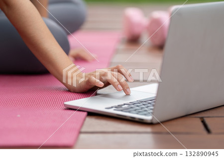 Remote Work and Fitness. Woman using laptop for online workout while seated on exercise mat. Remote Work and Fitness. Woman using laptop for online workout while seated on exercise mat. 132890945
