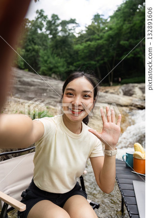 Camping and Joyful Moments. Woman waving at the camera while sitting by the river. Camping and Joyful Moments. Woman waving at the camera while sitting by the river. 132891606