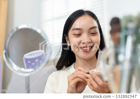 Makeup and Skincare Routine. A woman applying makeup while smiling at her reflection in a mirror. Makeup and Skincare Routine. A woman applying makeup while smiling at her reflection in a mirror. 132892347