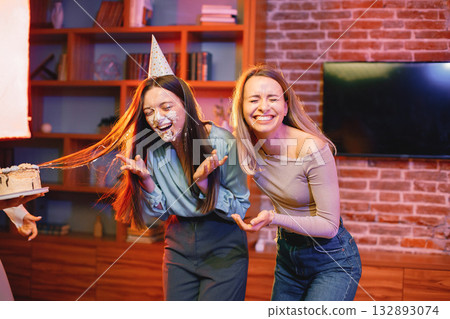 Young woman dips face in white cake with cream. Three girls having fun together. Birthday face cake smashing tradition concept. Young woman dips face in white cake with cream. Three girls having fun together. Birthday face cake smashing tradition concept. 132893074