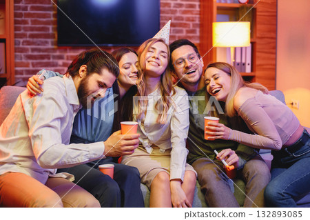 Young friends sitting on a coach in evening party red light. Blonde girl in a middle has a birthday and wearing party cone. Friends holding a paper cups with beverages.. 132893085