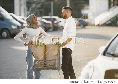 African Arabian couple stands with groceries near electric car. Charging electric car at the electric gas station African Arabian couple stands with groceries near electric car. Charging electric car at the electric gas station 132893092