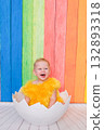 A young child is sitting in a white eggshell, wearing a yellow outfit 132893318