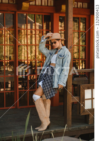 Stylish young woman poses confidently outside wooden building wearing denim jacket and wide-brim hat with sunglasses during sunny day in rustic setting Stylish young woman poses confidently outside wooden building wearing denim jacket and wide-brim hat with sunglasses during sunny day in rustic setting 132900413