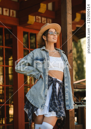 Stylish young woman poses confidently in a sunlit outdoor setting wearing denim, a hat, and sunglasses during a warm afternoon Stylish young woman poses confidently in a sunlit outdoor setting wearing denim, a hat, and sunglasses during a warm afternoon 132900414