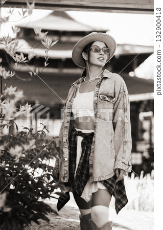 Stylish woman in denim jacket and hat stands confidently in a vibrant outdoor setting with flowers and traditional architecture in the background during daylight Stylish woman in denim jacket and hat stands confidently in a vibrant outdoor setting with flowers and traditional architecture in the background during daylight 132900418