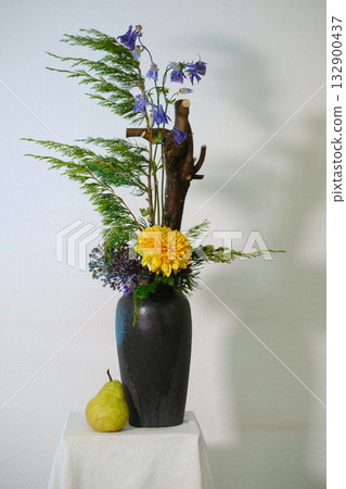 Elegant flower arrangement featuring yellow and purple blooms in a black vase with natural elements and a pear placed beside it on a white surface Elegant flower arrangement featuring yellow and purple blooms in a black vase with natural elements and a pear placed beside it on a white surface 132900437