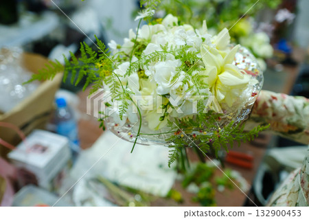 Beautiful floral arrangement featuring white flowers and greenery prepared for an event in a busy workspace during the afternoon Beautiful floral arrangement featuring white flowers and greenery prepared for an event in a busy workspace during the afternoon 132900453