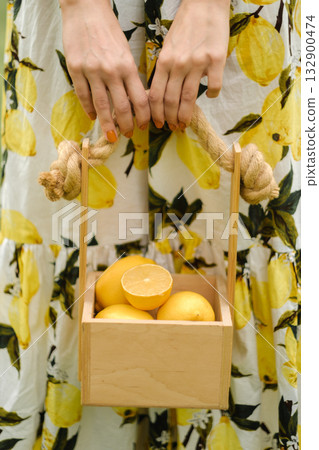 Bright yellow lemons in a wooden basket held by a person wearing a floral dress in a sunny outdoor setting, creating a fresh and vibrant atmosphere Bright yellow lemons in a wooden basket held by a person wearing a floral dress in a sunny outdoor setting, creating a fresh and vibrant atmosphere 132900474