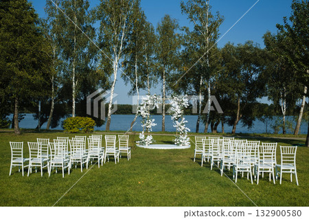 Beautiful outdoor wedding setup near a serene lake with white chairs arranged around a floral arch on a clear sunny day 132900580