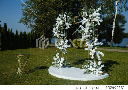 Elegant floral arch set up for a wedding ceremony by the lake in a tranquil park during a sunny afternoon Elegant floral arch set up for a wedding ceremony by the lake in a tranquil park during a sunny afternoon 132900581