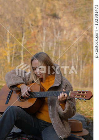 Beautiful blonde girl playing acoustic guitar and singing relaxing enjoying outdoors on sunny autumn day. Hippie woman in boho style learning and practicing playing musical instrument. Fall leaves 132901473