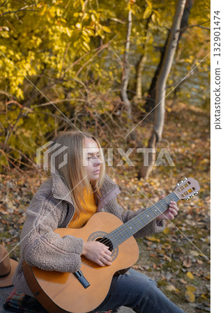 Blonde woman playing string guitar outdoors in autumn forest. Concept of sound therapy, mental health and wellness rituals. Calmness tranquility audio-sensory practices. Aura farming energy 132901474