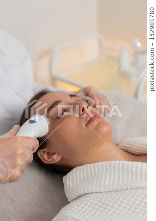 Beautician makes woman RF lifting cosmetology procedure of face and cheek area, collagen mask at spa Beautician makes woman RF lifting cosmetology procedure of face and cheek area, collagen mask at spa 132901780